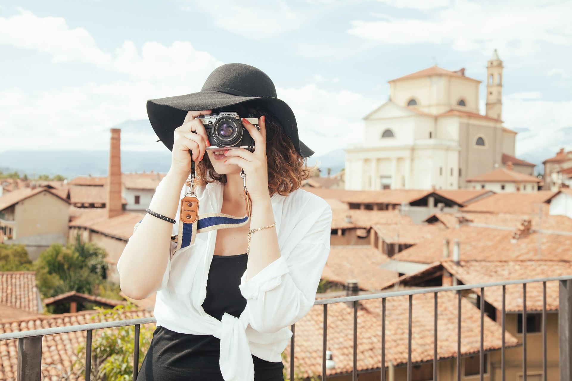 Eine Frau mit Hut steht auf einem Balkon und blickt durch eine Kamera.